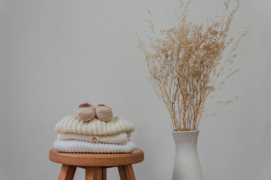 Folded knit baby sweaters and booties on a wooden stool beside dried flowers
